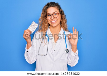 Young beautiful blonde doctor woman with blue eyes wearing stethoscope holding pills doing ok sign with fingers, excellent symbol