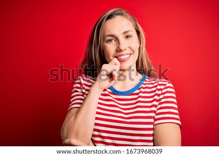 Young beautiful blonde woman wearing casual striped t-shirt over isolated red background looking confident at the camera with smile with crossed arms and hand raised on chin. Thinking positive.