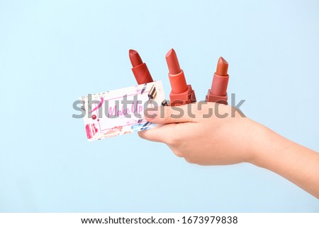 Female hand with business card of makeup artist and lipsticks on color background