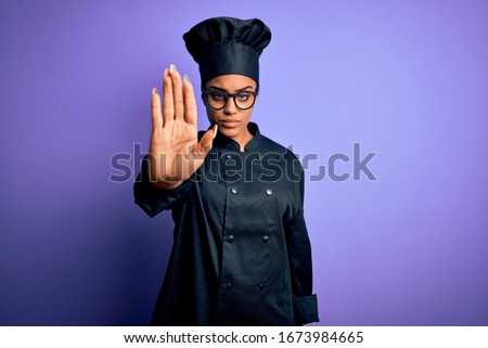 Young african american chef girl wearing cooker uniform and hat over purple background doing stop sing with palm of the hand. Warning expression with negative and serious gesture on the face.