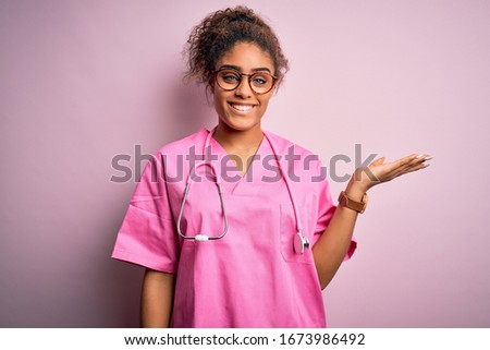 African american nurse girl wearing medical uniform and stethoscope over pink background smiling cheerful presenting and pointing with palm of hand looking at the camera.