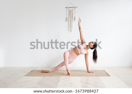 Young attractive woman practicing yoga in bright yoga class on wooden eco floors tanding in Full Version of Vasisthasana exercise, Side Plank pose, working out, pink sportswear. White wall background Royalty-Free Stock Photo #1673988427