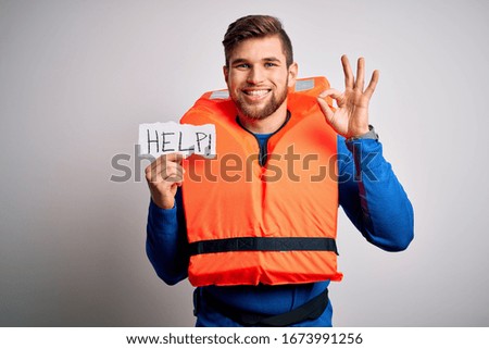 Young blond man with beard and blue eyes wearing lifejacket holding paper with help message doing ok sign with fingers, excellent symbol
