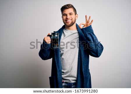 Young blond man with beard and blue eyes wearing pajama making coffe using coffeemaker doing ok sign with fingers, excellent symbol