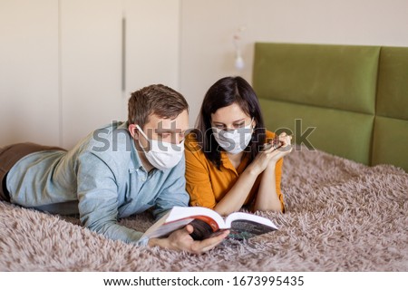 A young couple is sitting at home on self-quarantine. Husband and wife quarantined  coronavirus in protective masks. New reality. Normal life in isolation. Read a book together. Italy europe covid-19 Royalty-Free Stock Photo #1673995435