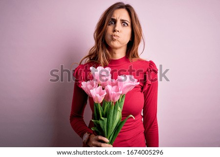 Young beautiful brunette woman holding bouquet of pink tulips over isolated background puffing cheeks with funny face. Mouth inflated with air, crazy expression.
