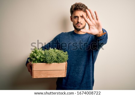 Young gardener man with beard holding box with plants standing over white background with open hand doing stop sign with serious and confident expression, defense gesture