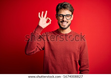 Young handsome man with beard wearing glasses and sweater standing over red background smiling positive doing ok sign with hand and fingers. Successful expression.