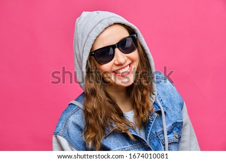 Stylish Caucasian girl in a blue jeans and 8-bit glasses and smiling at the camera on a pink background. Close up.
