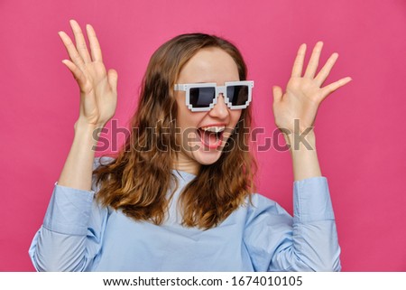 Stylish caucasian girl in a pale blue t-shirt and 8-bit glasses waving arms to the top on a pink background. Close up.