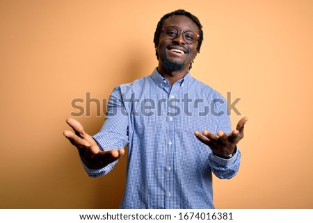 Young handsome african american man wearing shirt and glasses over yellow background smiling cheerful offering hands giving assistance and acceptance.
