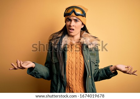 Young beautiful hispanic woman wearing ski glasses and coat for winter weather clueless and confused with open arms, no idea concept.