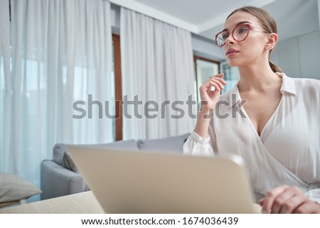 woman in eyeglasses sitting with computer on couch, looking outside, cannot concentrate on work, need some rest, feeling bored, need additional motivation, working remotely at home.