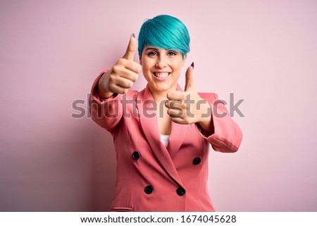 Young beautiful businesswoman with blue fashion hair wearing jacket over pink background approving doing positive gesture with hand, thumbs up smiling and happy for success. Winner gesture.