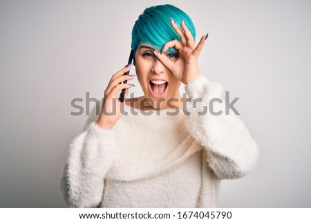 Young woman with blue fashion hair having a conversation talking on smartphone with happy face smiling doing ok sign with hand on eye looking through fingers