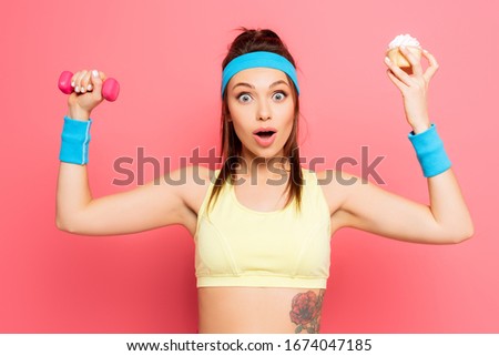 shocked young sportswoman holding dumbbell and delicious cupcake while looking at camera on pink background
