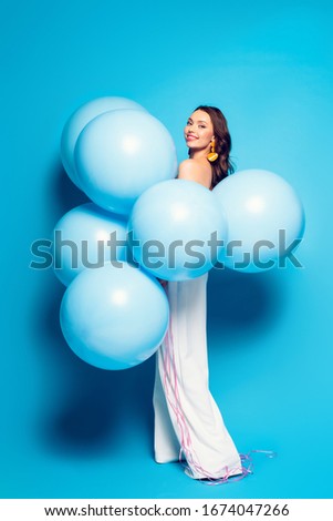 full length view of happy elegant woman with big festive balloons smiling at camera on blue background