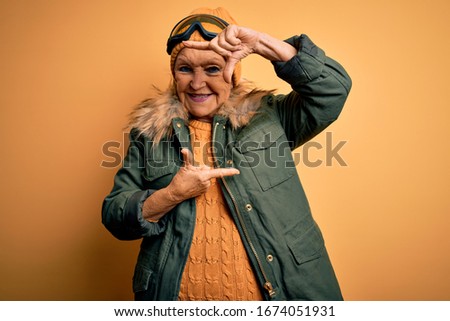 Senior beautiful grey-haired skier woman wearing snow sportswear and ski goggles smiling making frame with hands and fingers with happy face. Creativity and photography concept.