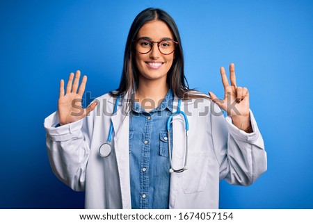 Young beautiful doctor woman wearing stethoscope and glasses over blue background showing and pointing up with fingers number eight while smiling confident and happy.