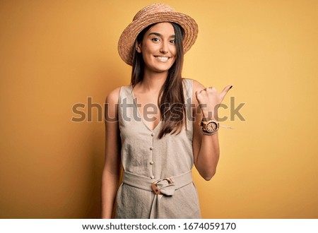 Young beautiful brunette woman on vacation wearing casual dress and hat smiling with happy face looking and pointing to the side with thumb up.