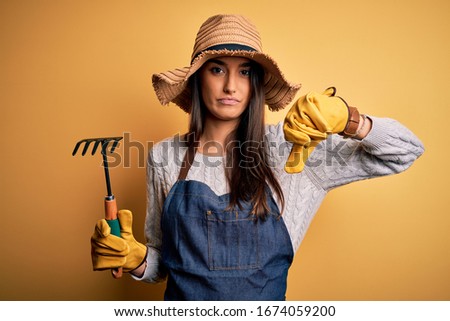 Young beautiful farmer woman wearing apron and hat using rake over yellow background with angry face, negative sign showing dislike with thumbs down, rejection concept