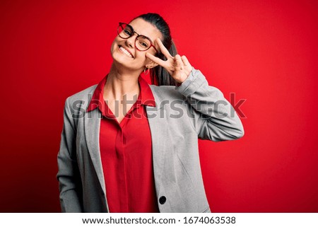 Young beautiful brunette businesswoman wearing jacket and glasses over red background Doing peace symbol with fingers over face, smiling cheerful showing victory