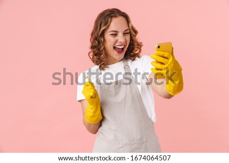 Image of a happy excited young housewife woman in gloves holding mobile phone and credit card isolated over pink wall background.