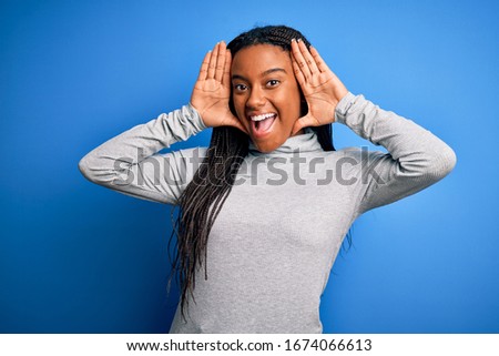 Young african american woman standing wearing casual turtleneck over blue isolated background Smiling cheerful playing peek a boo with hands showing face. Surprised and exited