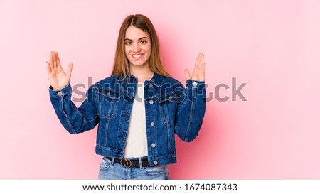 Young caucasian woman isolated on pink background holding something little with forefingers, smiling and confident.