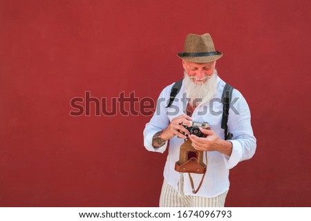 Happy hipster man using old vintage camera standing on red wall - Mature stylish photographer having fun traveling the world - Joyful elderly lifestyle concept - Focus on face