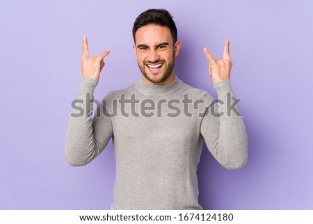 Young caucasian man isolated on purple background showing a horns gesture as a revolution concept.