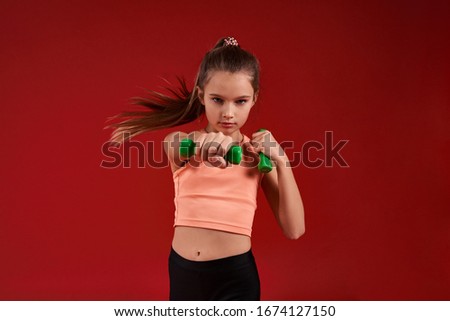 A cute kid, girl is engaged in sport, she is looking at camera while exercising with dumbbells. Isolated on red background. Fitness, training, active lifestyle concept. Horizontal shot