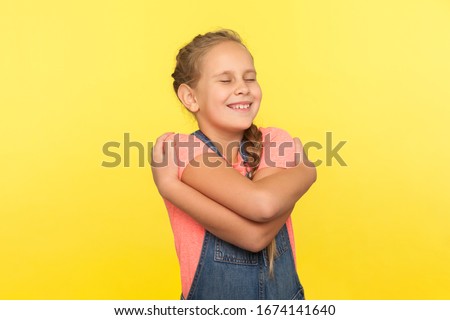 Portrait of happy cute little girl with braid in denim overalls embracing herself and smiling, concept of self-love, positive self-esteem in childhood. indoor studio shot isolated on yellow background