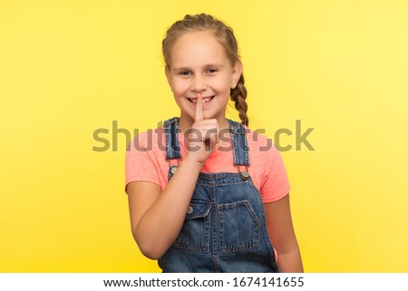 Be quiet, it's secret. Portrait of positive little girl with braid in denim overalls making silence gesture with finger on lips, child mystery. indoor studio shot isolated on yellow background