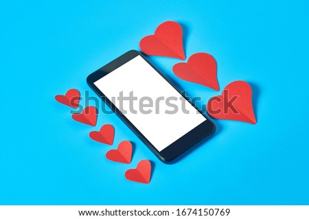 Black smartphone with isolated white screen for text, picture, photo and other graphics near rows of red paper hearts on blue table. Valentines day and love concept. Space for text