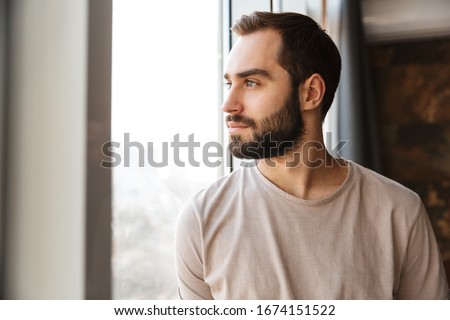 Handsome serious young bearded man standing at the window at home, looking away Royalty-Free Stock Photo #1674151522
