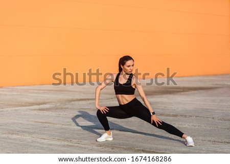 Slim young pretty woman in tight sportswear, black pants and top, practicing outdoor, doing stretching workouts for better flexibility, lower-body exercise. Health care concept, sport activity Royalty-Free Stock Photo #1674168286