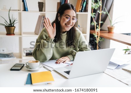 Attractive happy young girl student studying at the college library, sitting at the desk, using laptop computer, having video chat, waving Royalty-Free Stock Photo #1674180091