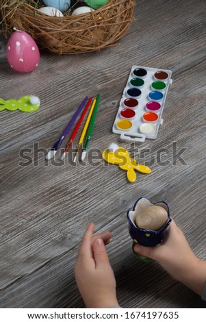 baby hand turn easter egg. In wooden table paints, brushes, bunny, nest. Prepares to paint Easte eggs