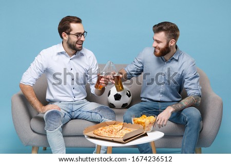 Funny two young men guys friends colleagues in casual shirt sit on couch isolated on pastel blue background. Sport leisure concept. Cheer up support favorite team with soccer ball hold beer bottle