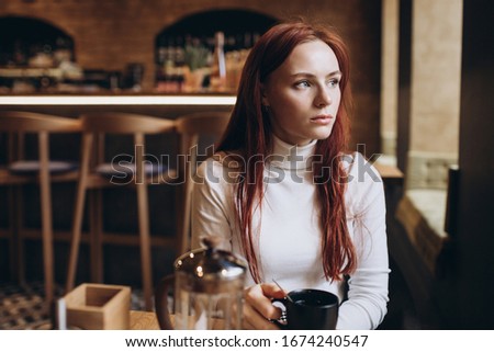 Close up portrait of young attractive ginger girl with freckles sitting i a cafe drinking coffe alone, looking in the window. Emotional concept, freckled people concept