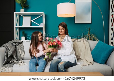 I love my mom! Attractive young woman with little cute girl are spending time together at home. Little daughter greeting mom with bouquet of tulips. Happy family concept. Mother's day