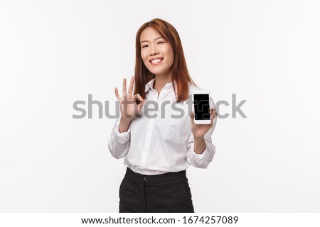 This is good. Satisfied good-looking asian woman showing mobile phone display and make okay gesture, rate excellent app, feeling proud of taking cool pic, standing white background