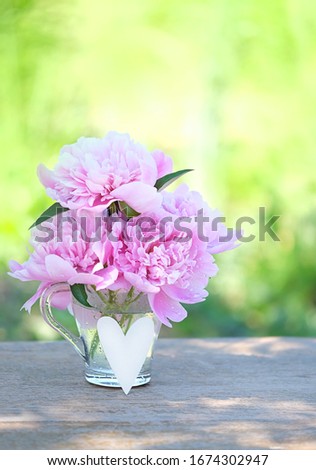beautiful Peony Flowers in glass cup and heart decor on table in garden, abstract natural background. gentle romantic composition with flowers. spring, summer season. template for design. copy space.