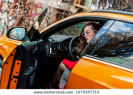 Beautiful girl in a red coat posing near in orange car. girl in a coat outdoors. beautiful girl in a fashionable casual coat. spring autumn collection.