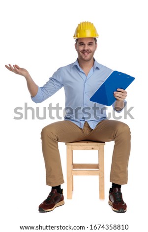 Positive casual man wearing hard hat inviting with clipboard in hand while sitting on a chair on white studio background