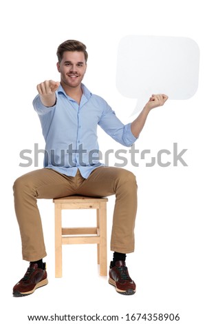 Happy casual man holding speech bubble and pointing forward, smiling while sitting on a chair on white studio background