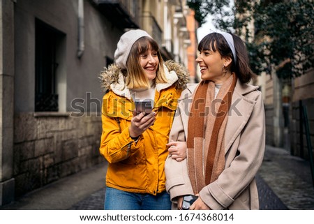 Stock photo of two caucasian girls walking in the street together laughing and having fun while using a smartphone. They are looking between them.