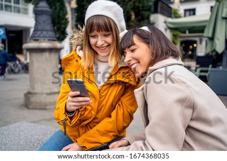 Stock photo of two caucasian girls looking at a smartphone. They are laughing. They are seated on the street.