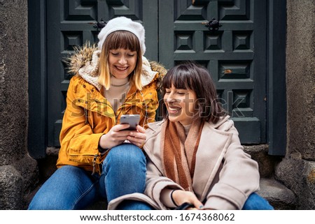 Stock photo of two caucasian girls looking at a smartphone. They are laughing. They are seated on the street.